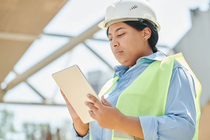 Profissional com capacete e colete de segurança consultando um tablet em área de obra